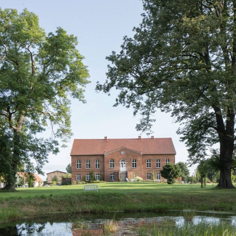 fasten-mit-diana/fastenwandern Zweistöckiges, rotes Backsteingebäude mit großen Fenstern und einem Garten davor.